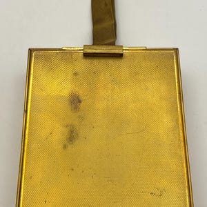 May include: A vintage gold-toned compact mirror with a textured surface and a matching fabric handle. The rectangular compact has a simple, elegant design, perfect for carrying in a purse or pocket. The compact is a classic accessory.