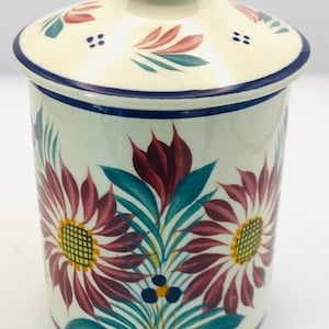 May include: A ceramic jar with a lid, featuring a floral design in burgundy, teal, and yellow on a cream background. The lid has a dark blue knob and trim. The jar's base and rim are also trimmed in dark blue.