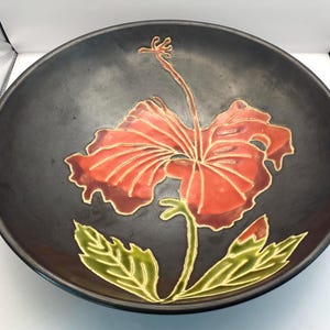 Op de afbeelding: Een zwarte keramische kom met een rood hibiscusbloemdesign. De bloem heeft gouden contouren en groene bladeren. De kom is rond en heeft een glad oppervlak.