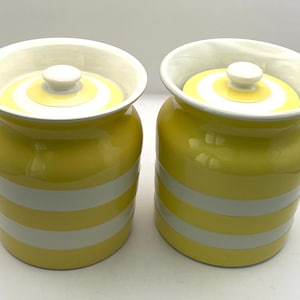 May include: Two yellow ceramic canisters with white horizontal stripes. Each canister has a white lid with a yellow center and a white knob. The canisters are likely for kitchen storage.