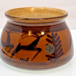 Könnte beinhalten: Eine runde, braune Keramikschale mit glänzender Oberfläche, verziert mit dunkelbraunen, handgemalten abstrakten Tier- und Baummotiven. Ein kleiner runder Aufkleber mit japanischem Text und dem Wort Lap-d ist an der Außenseite der Schale angebracht.