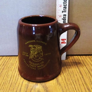Może przedstawiać: Brązowy ceramiczny kubek z uchwytem. Kubek ma złoty tekst i grafikę, w tym "MACOMB, ILLINOIS", "OLD SLEEPY EYE" i "FOURTH ANNUAL COLLECTORS CONVENTION" z datami "JULY 27-28 1979."