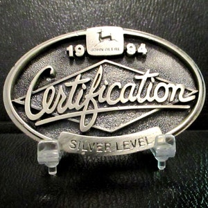 May include: Silver metal belt buckle with the text "Certification" and "Silver Level" and the John Deere logo. The buckle also has the year "1984" on it.