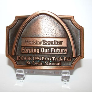 May include: A bronze-coloured belt buckle featuring a depiction of the St. Louis Arch. The buckle has the text "Working Together Forging Our Future JI CASE 1994 Parts Trade Fair St. Louis, Missouri".