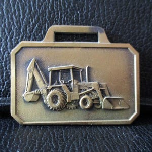 Könnte beinhalten: Rechteckiger, bronzefarbener Metallanhänger mit Griff. Detailliertes Relief eines Baggerladers. Der Anhänger hat eine strukturierte Oberfläche. Der Baggerlader ist mit ausgefahrenem Löffel und Arm dargestellt.