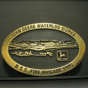 May include: A gold-colored oval belt buckle with the text "John Deere Waterloo Works D.S.S. Fire Brigade 1995" and a black and white image of a factory. The John Deere logo is in the lower right corner.