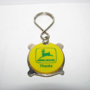 May include: A yellow and silver keyring with a green John Deere logo and the word "Thanks". The logo features a green deer with antlers.