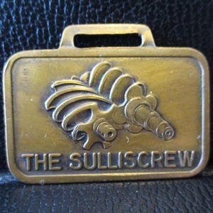 Puede incluir: Un llavero de latón con una imagen estilizada de un tornillo y el texto "THE SULLISCREW".