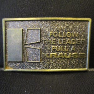 May include: Rectangular brass belt buckle with the text "FOLLOW THE LEADER PULL A KRAUSE". The buckle has a textured background and a silver-colored design on the left side. The edges are framed with a gold border.