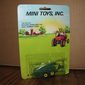 May include: A green 1/64 scale toy manure spreader with real rubber tires. The toy is packaged on a card with a cartoon illustration of a red tractor in a field.