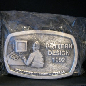 May include: A silver belt buckle with a design of a person working on a computer. The buckle says "Pattern Design 1992" and "John Deere Foundry Waterloo".