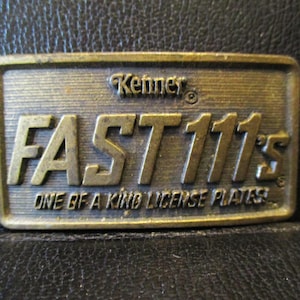 May include: A rectangular, brass-colored metal belt buckle with the text "Kenner FAST 111's ONE OF A KIND LICENSE PLATES" embossed on it. The buckle has a vintage, worn appearance.