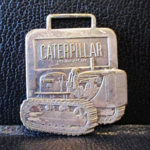 May include: A silver-coloured Caterpillar tractor pin featuring the word "CATERPILLAR". The pin has a rectangular shape with a handle at the top and a detailed tractor design.