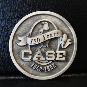 Puede incluir: Una moneda conmemorativa de color plateado con el texto "150 Years" en una pancarta, y "CASE 1842-1992" debajo. Un águila está centrada dentro del diseño de la moneda. La moneda está sobre un fondo negro texturizado.
