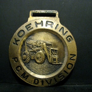 以下が含まれることがあります： 縁に「Koehring PCM Division」と刻まれた金色の金属製のキーホルダー。中央には建設車両の小さな詳細な画像があります。
