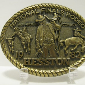 Op de afbeelding: Een gouden metalen gesp met een touwachtige rand. De gesp toont een cowboy die een wild paard berijdt, een cowboy die een kalf met een lasso vangt en een cowboy die een lasso vasthoudt. De gesp heeft ook de tekst "National Finals Rodeo", "All Around Cowboy", "19", "Hesston" en "82".