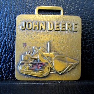 Op de afbeelding: Een gouden metalen sleutelhanger met een John Deere tractor met een schep. De sleutelhanger heeft een lus aan de bovenkant om aan sleutels te bevestigen.