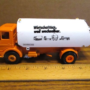 Puede incluir: Camión de juguete naranja y blanco con una caja blanca en la parte trasera. La caja tiene el texto "Wirtschaftlich, weil wechselbar." y una serie de ilustraciones en blanco y negro del camión con diferentes accesorios. El camión mide aproximadamente 12,7 cm de largo.