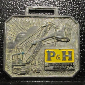 Peut inclure: Un porte-clé de montre de poche en métal argenté avec une image en noir et blanc d'une excavatrice avec un logo P&H jaune. Le logo indique "Quality & Service Since 1928".