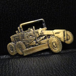 May include: A brass-toned metal pin shaped like a John Deere motor grader. The construction vehicle features a detailed cab, a central blade, and multiple wheels. The surface has a weathered, antique finish with dark recessed areas highlighting the mechanical components of the heavy machinery.