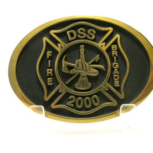 May include: A gold-colored metal belt buckle with a black enamel center. The center features a fire department logo with a crossed axe and hose, a fireman's helmet, and the text "DSS FIRE BRIGADE 2000".