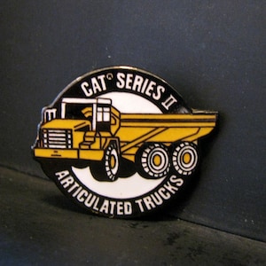 May include: A yellow and black pin featuring a cartoon illustration of a Caterpillar articulated truck with the text "CAT Series II Articulated Trucks".