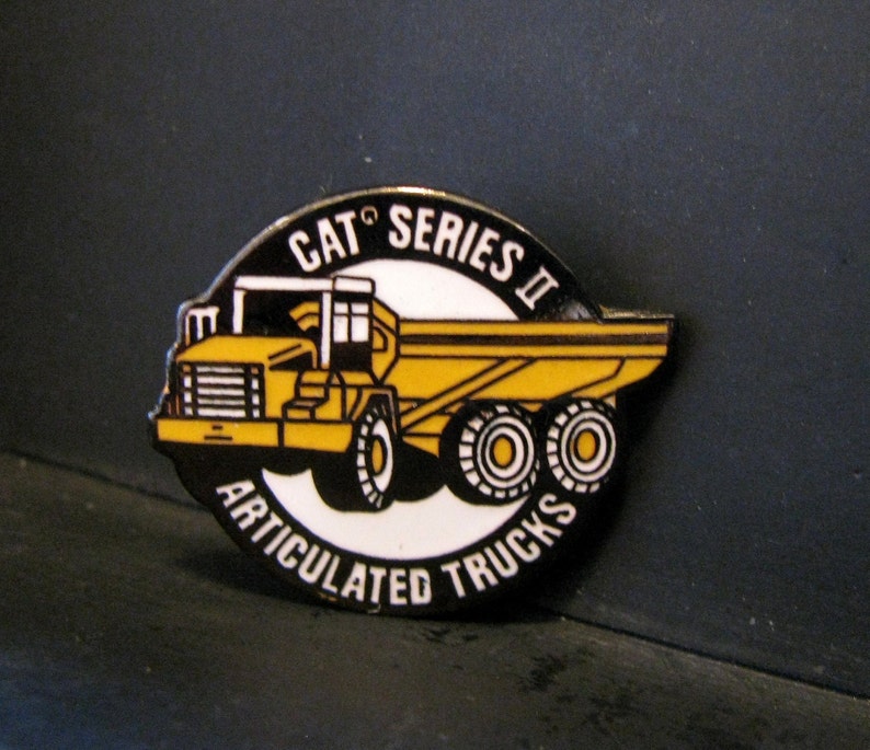 Op de afbeelding: Een gele en zwarte pin met een cartoonillustratie van een gelede Caterpillar-vrachtwagen met de tekst "CAT Series II Articulated Trucks".