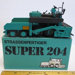 Puede incluir: Un modelo de juguete de pavimentadora vial, de color verde azulado y negro, modelo Super 204, sobre su caja original. La caja es verde azulado con texto negro que dice "STRASSENFERTIGER SUPER 204". El juguete tiene múltiples ruedas y un diseño detallado.
