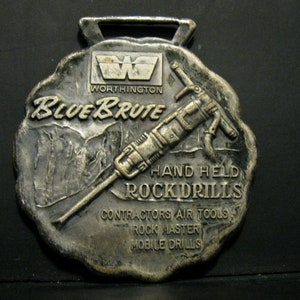 Puede incluir: Un medallón de metal plateado con una imagen en relieve de un martillo neumático y el texto "Blue Brute" y "Worthington". También se puede ver el texto "Hand Held Rock Drills", "Contractors Air Tools", "Rock Master" y "Mobile Drills".