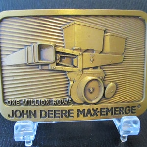 May include: A gold-colored metal belt buckle featuring a 3D image of a John Deere Max-Emerg planter with the text "One Million Rows" and "John Deere Max-Emerg".
