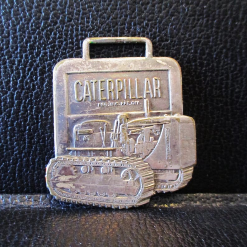 May include: A silver-coloured Caterpillar tractor pin featuring the word "CATERPILLAR". The pin has a rectangular shape with a handle at the top and a detailed tractor design.