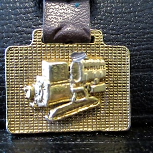 May include: Gold-colored metal keychain featuring a detailed model of a small engine with the word "Homelite" embossed on the side.