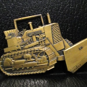 Op de afbeelding: Een gouden metalen gesp met een gedetailleerd model van een John Deere 610 bulldozer. De bulldozer heeft een reliëf, textuurontwerp en is geplaatst tegen een zwarte achtergrond.