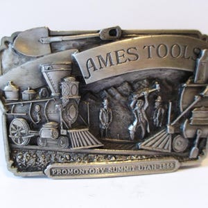 May include: A silver-toned metal belt buckle featuring a detailed relief of a steam train, workers, and the text "AMES TOOLS" and "PROMONTORY SUMMIT UTAH 1869". The design includes a shovel and a banner.