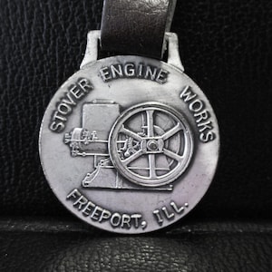 Puede incluir: Un llavero de reloj de bolsillo plateado con una correa de cuero negro. El llavero presenta una imagen de un motor antiguo con el texto "Stover Engine Works Freeport, Ill."