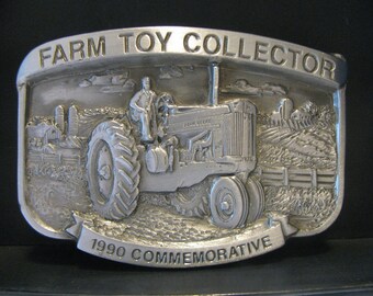 May include: Silver belt buckle with a 1990 commemorative inscription. The buckle features a detailed image of a John Deere tractor in a farm setting.