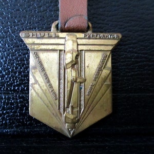 May include: A gold-coloured metal badge with the words "Shull Perforator" and "Knife Perforator" on it. The badge also has the words "Cut-Away View of Long" on it.