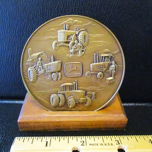 Puede incluir: Una medalla de bronce con cuatro tractores John Deere en relieve. La medalla está sobre un soporte de madera. La medalla tiene el logotipo de John Deere y las palabras "John Deere" en ella.