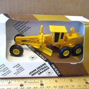 Puede incluir: Un juguete de niveladora industrial John Deere amarillo a escala 1/64 en su caja original. La caja presenta una descripción detallada de las características del juguete, incluyendo un eje delantero pivotante, dirección articulada, una cuchilla giratoria y una construcción de metal fundido a presión duradera.