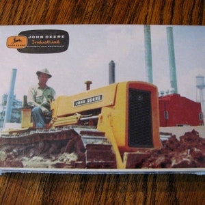 May include: A yellow John Deere Industrial tractor with a man in a white hat sitting in the driver's seat. The tractor is in front of a red building with tall smokestacks in the background.