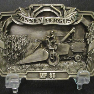 May include: Silver-toned belt buckle featuring a 3D image of a Massey Ferguson MF 35 combine harvester in a field of corn. The buckle has a banner with the words "Massey Ferguson" above the combine.