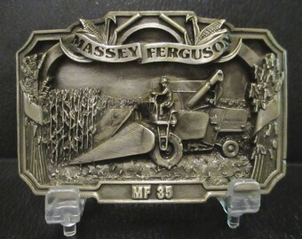 May include: Silver-toned belt buckle featuring a 3D image of a Massey Ferguson MF 35 combine harvester in a field of corn. The buckle has a banner with the words "Massey Ferguson" above the combine.