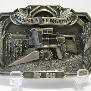 May include: A silver-toned belt buckle featuring a detailed 3D image of a Massey Ferguson MF 540 combine harvester in a field of wheat. The buckle has a banner with the words "Massey Ferguson" and the model number "MF 540" is inscribed below the harvester.
