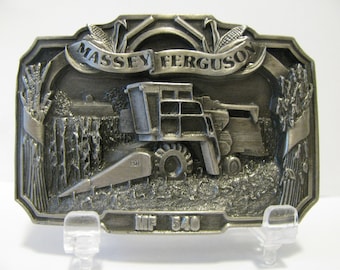 May include: A silver-toned belt buckle featuring a detailed 3D image of a Massey Ferguson MF 540 combine harvester in a field of wheat. The buckle has a banner with the words "Massey Ferguson" and the model number "MF 540" is inscribed below the harvester.