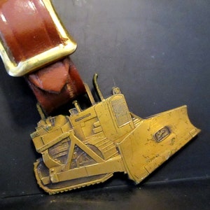 Op de afbeelding: Een gouden metalen gesp met een gedetailleerd model van een Caterpillar bulldozer. De bulldozer heeft een bruine leren riem.