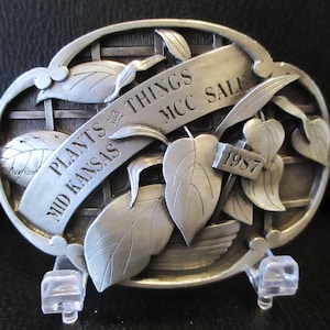May include: Silver-toned metal belt buckle with a detailed design of a plant with leaves and a banner that reads "Plants & Things Mid Kansas MCC Sale 1987".