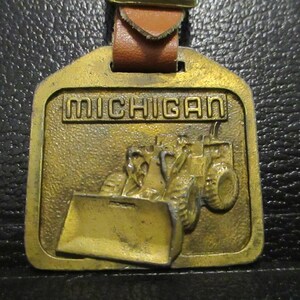 Peut inclure: Un porte-clés en métal doré avec une lanière en cuir marron. Le porte-clés représente un tracteur Michigan avec une lame en relief. Le texte "MICHIGAN" est en relief sur le porte-clés.