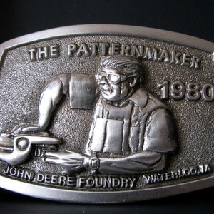 Può includere: Fibbia per cintura in argento con un'immagine in rilievo di un uomo che indossa occhiali di sicurezza e una camicia da lavoro. La fibbia riporta la scritta "The Patternmaker 1980 John Deere Foundry Waterloo, IA".