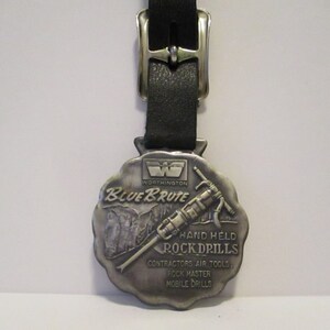 Puede incluir: Un llavero de metal plateado con un logotipo de Worthington Blue Brute y una representación de un martillo neumático. El llavero dice "Hand Held Rock Drills, Contractors Air Tools, Rock Master Mobile Drills".