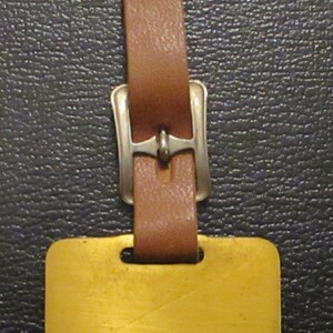 May include: A brown leather luggage tag with a silver buckle and a blank gold metal tag.